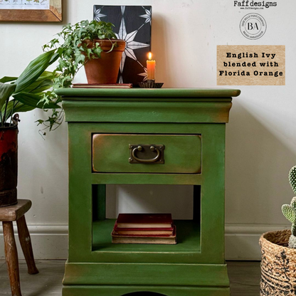Green wooden side table with a drawer, books, and potted plants in a room setting.