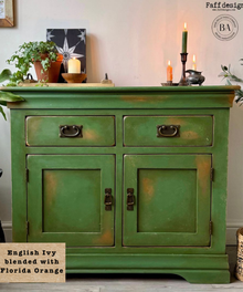 A vintage-style wooden sideboard cabinet painted in a distressed green finish with dark metal drawer and door handles, shown in a home interior setting with candles, a plant, and framed art.
