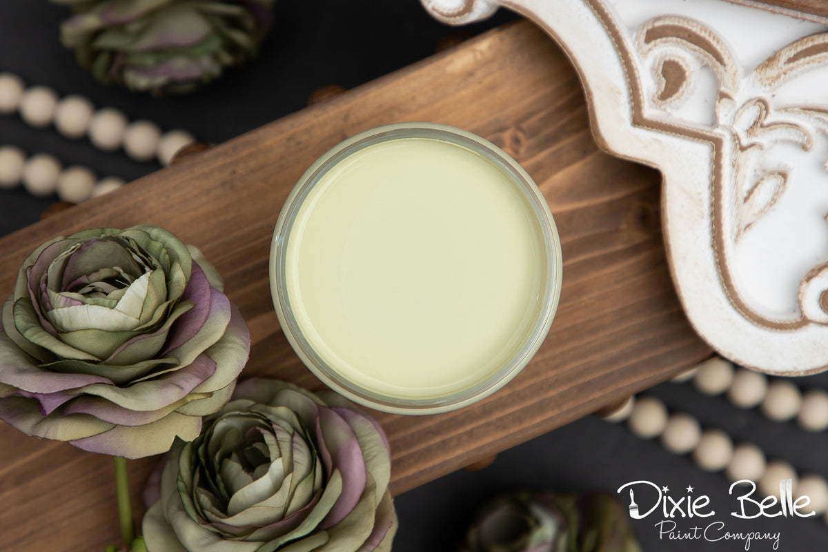 An open jar of Dixie Belle Paint Company farmhouse green colored paint sits on a wooden board, surrounded by decorative faux roses and beads.