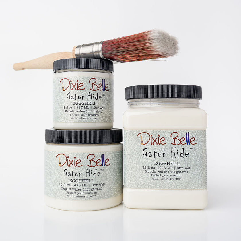 Three jars of Dixie Belle Gator Hide eggshell acrylic paint glazes with a paintbrush resting on top, arranged on a clean white background.