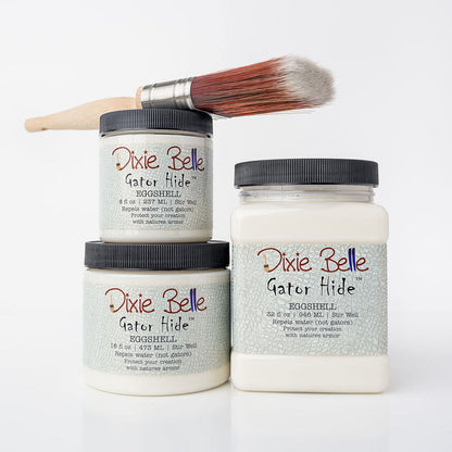 Three jars of Dixie Belle Gator Hide eggshell acrylic paint glazes with a paintbrush resting on top, arranged on a clean white background.