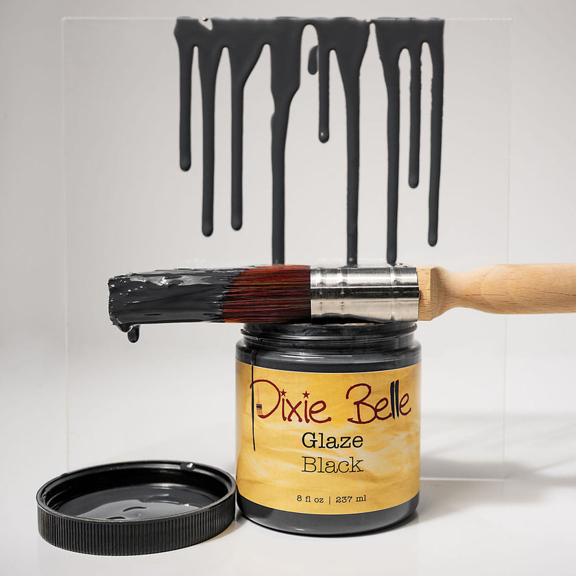 A jar of Dixie Belle Glaze Black paint with its lid off, a paintbrush coated in black paint resting across the jar, and black paint dripping from a clear panel above on a white background.