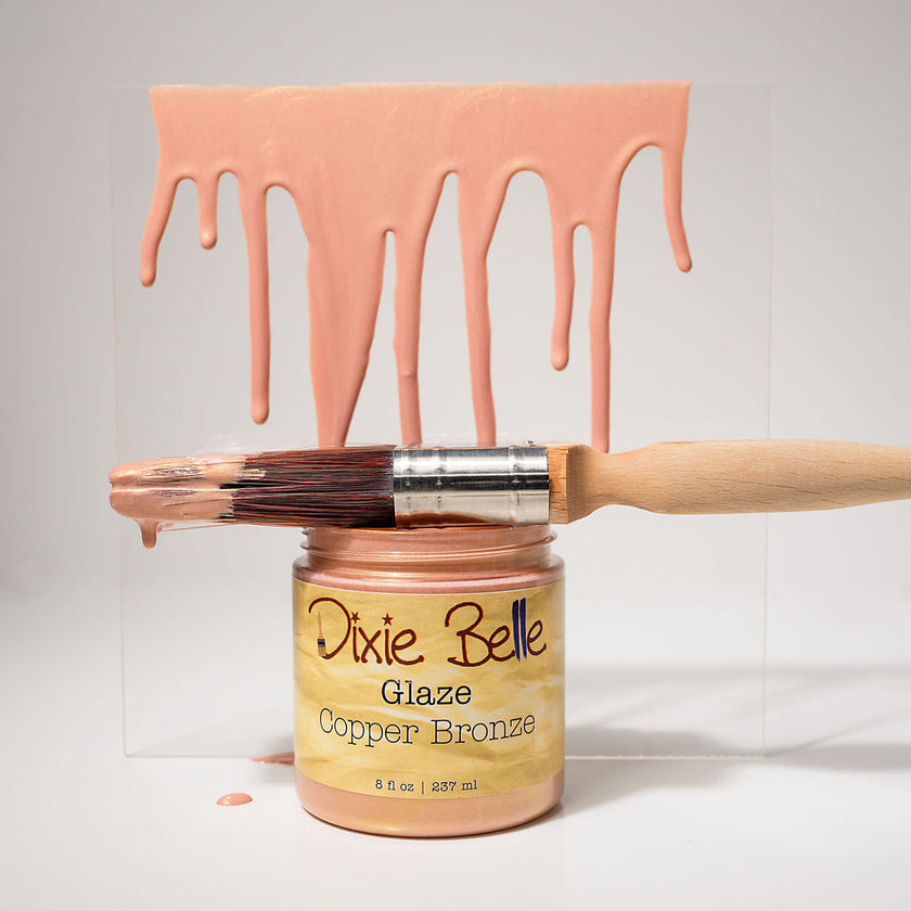 A jar of Dixie Belle Glaze Copper Bronze paint glaze with a wooden-handled brush resting across the open jar, set against a white background with a clear acrylic panel behind it. Pinkish copper-bronze paint is shown dripping from the top of the panel and also from the brush bristles.