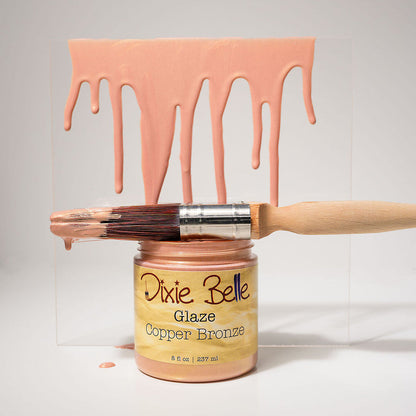 A jar of Dixie Belle Glaze Copper Bronze paint glaze with a wooden-handled brush resting across the open jar, set against a white background with a clear acrylic panel behind it. Pinkish copper-bronze paint is shown dripping from the top of the panel and also from the brush bristles.