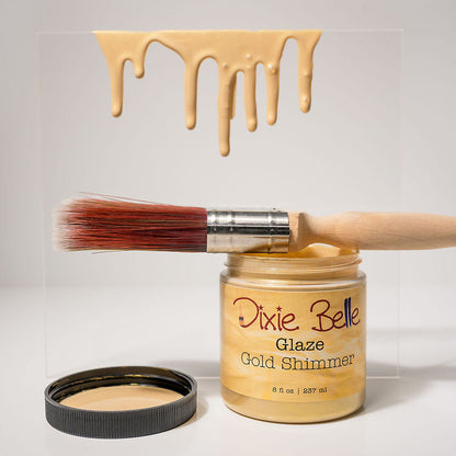 A jar of Dixie Belle Glaze Gold Shimmer nail polish with its lid off, a wooden-handled brush resting across the open jar, and a clear square card showing the gold shimmer polish dripping down, all on a light gray background.