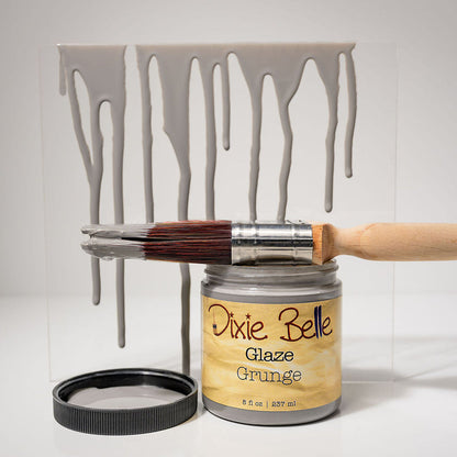 A jar of Dixie Belle Glaze Grunge paint with a paintbrush resting across the open jar and a black lid beside it, set in front of a clear panel showing gray paint dripping vertically. The background is a clean white studio backdrop.
