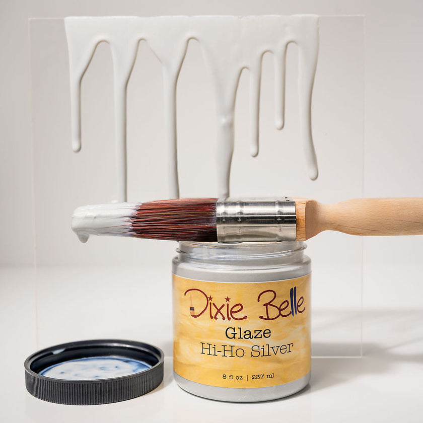 A jar of Dixie Belle Hi-Ho Silver Glaze paint with a paintbrush resting across the open jar and a lid placed beside it, set against a white background with a painted drip effect behind it.