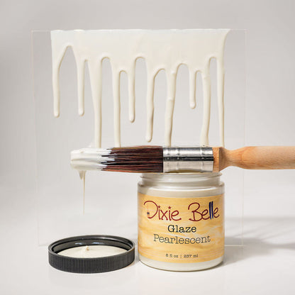 A jar of Dixie Belle Glaze Pearlescent paint with a wooden-handled brush resting across the open lid, and a clear acrylic panel showing a dripping white paint effect. The background is a clean light gray studio backdrop.