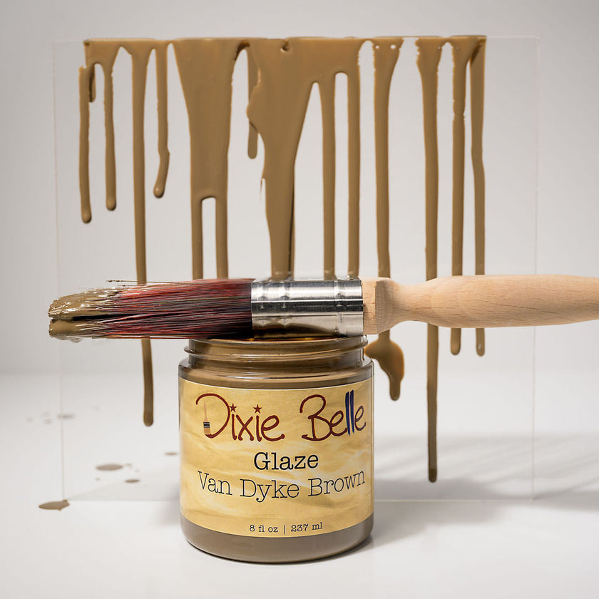 A jar of Dixie Belle Glaze Van Dyke Brown paint with a paintbrush resting across the open jar, set against a white background with a clear panel showing dripping paint in the same color.