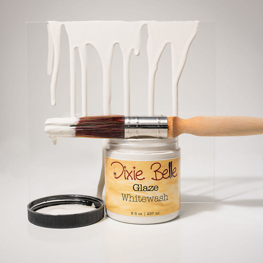 A jar of Dixie Belle Glaze Whitewash paint with a wooden-handled brush resting across the open jar and a black lid beside it, set against a white background with a clear panel showing dripping white paint.