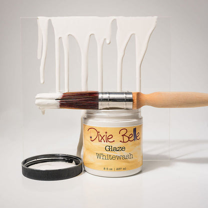 A jar of Dixie Belle Glaze Whitewash paint with a wooden-handled brush resting across the open jar and a black lid beside it, set against a white background with a clear panel showing dripping white paint.