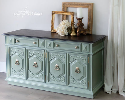 A painted sideboard console table with ornate carved doors, silver-tone drawer pulls and cabinet handles, a dark wood top, and decorative picture frames and flowers on top. The image is taken in a styled room setting with a curtain on the right.