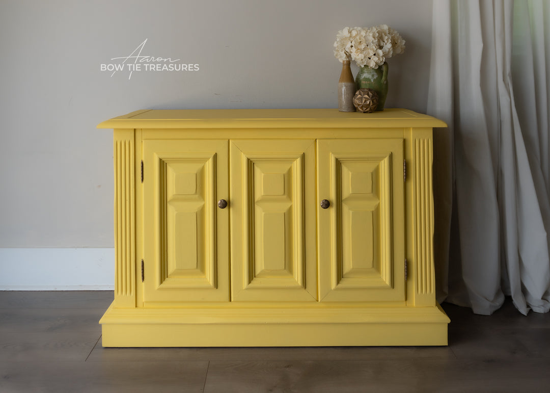 A bright yellow painted wooden sideboard cabinet with three paneled doors and small round knobs, shown in a styled room setting with a vase of flowers on top and a curtain to the right.