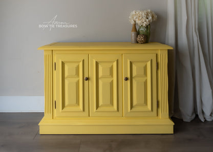A bright yellow painted wooden sideboard cabinet with three paneled doors and small round knobs, shown in a styled room setting with a vase of flowers on top and a curtain to the right.