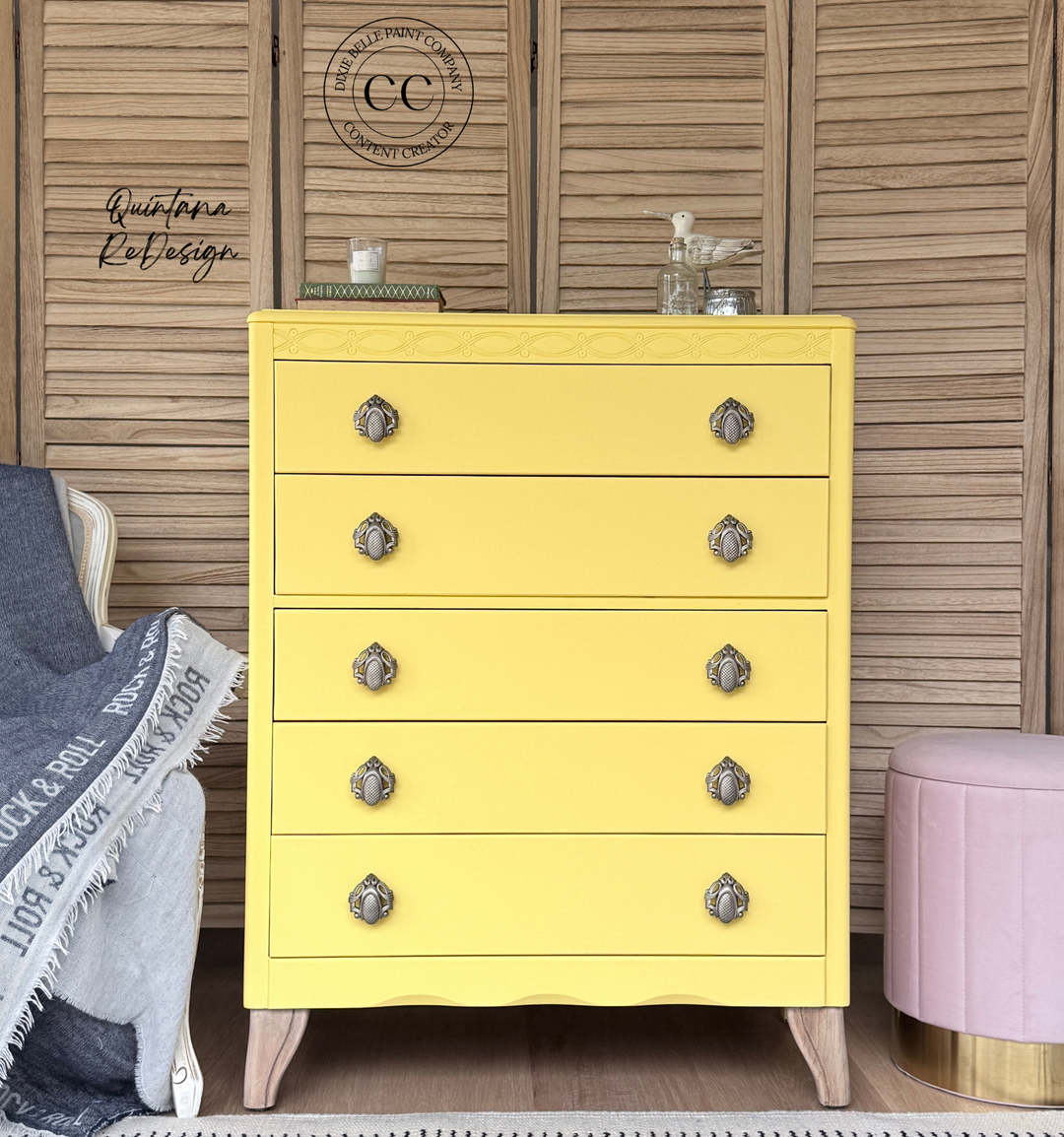 A bright yellow five-drawer chest of drawers with ornate silver knobs, styled in a room setting against wooden shutters with a pink ottoman and a throw blanket nearby.