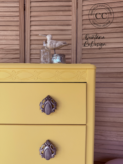 A close-up of a yellow painted dresser with ornate dark metal drawer pulls, set against a brown wooden slatted wall. A small bird figurine and glass jar sit on top.