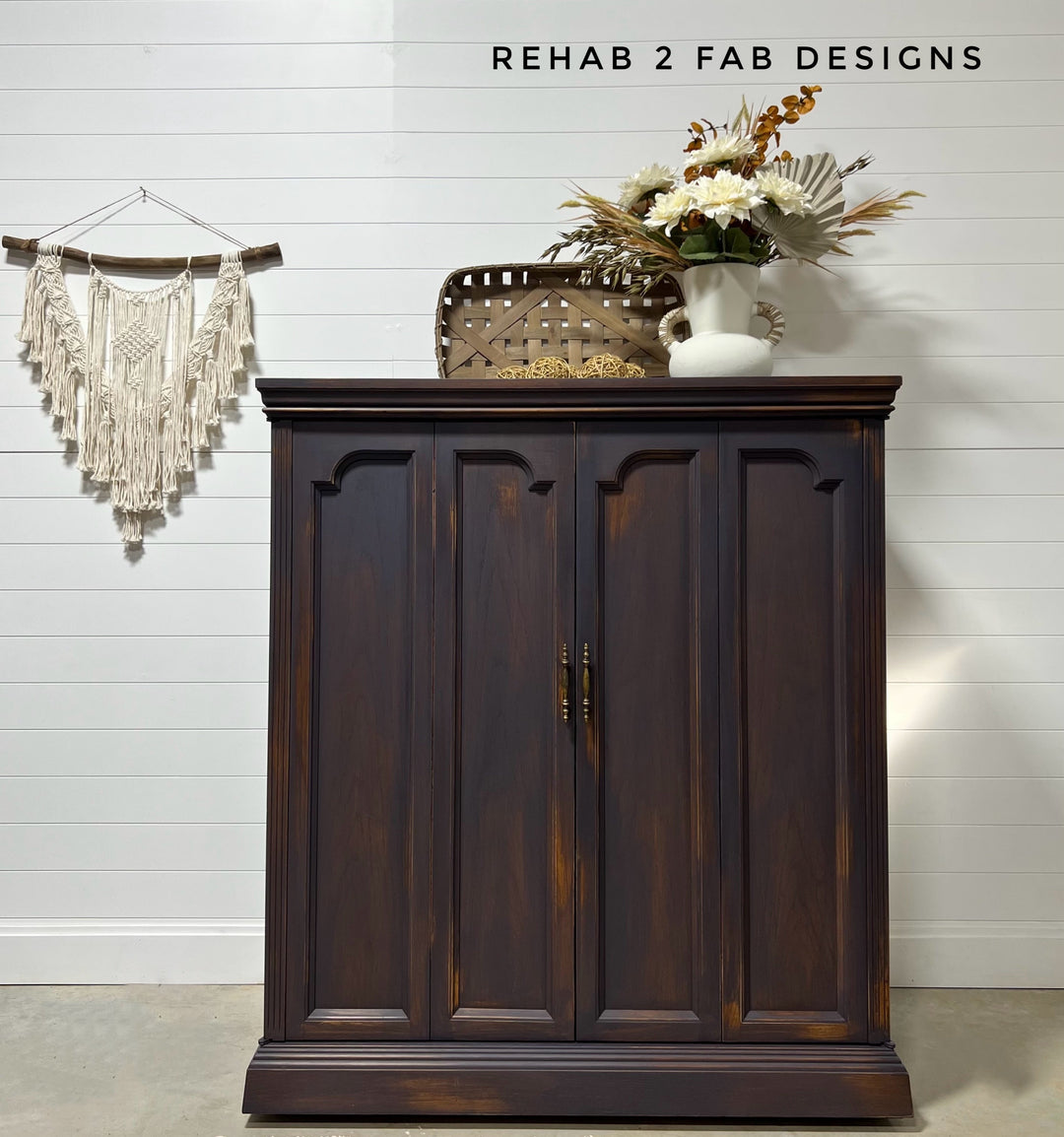 A dark brown wooden cabinet with three paneled doors and brass handles, styled with a woven basket, a white vase of dried flowers, and a macrame wall hanging against a white shiplap wall. Text at the top reads 'REHAB 2 FAB DESIGNS'.