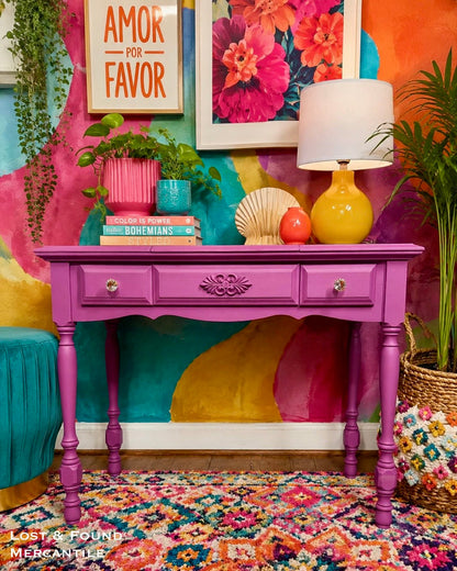 A bright magenta purple wooden console table styled with a yellow lamp, pink and blue ceramic pots, books, and decorative objects, set against a highly colorful painted wall.