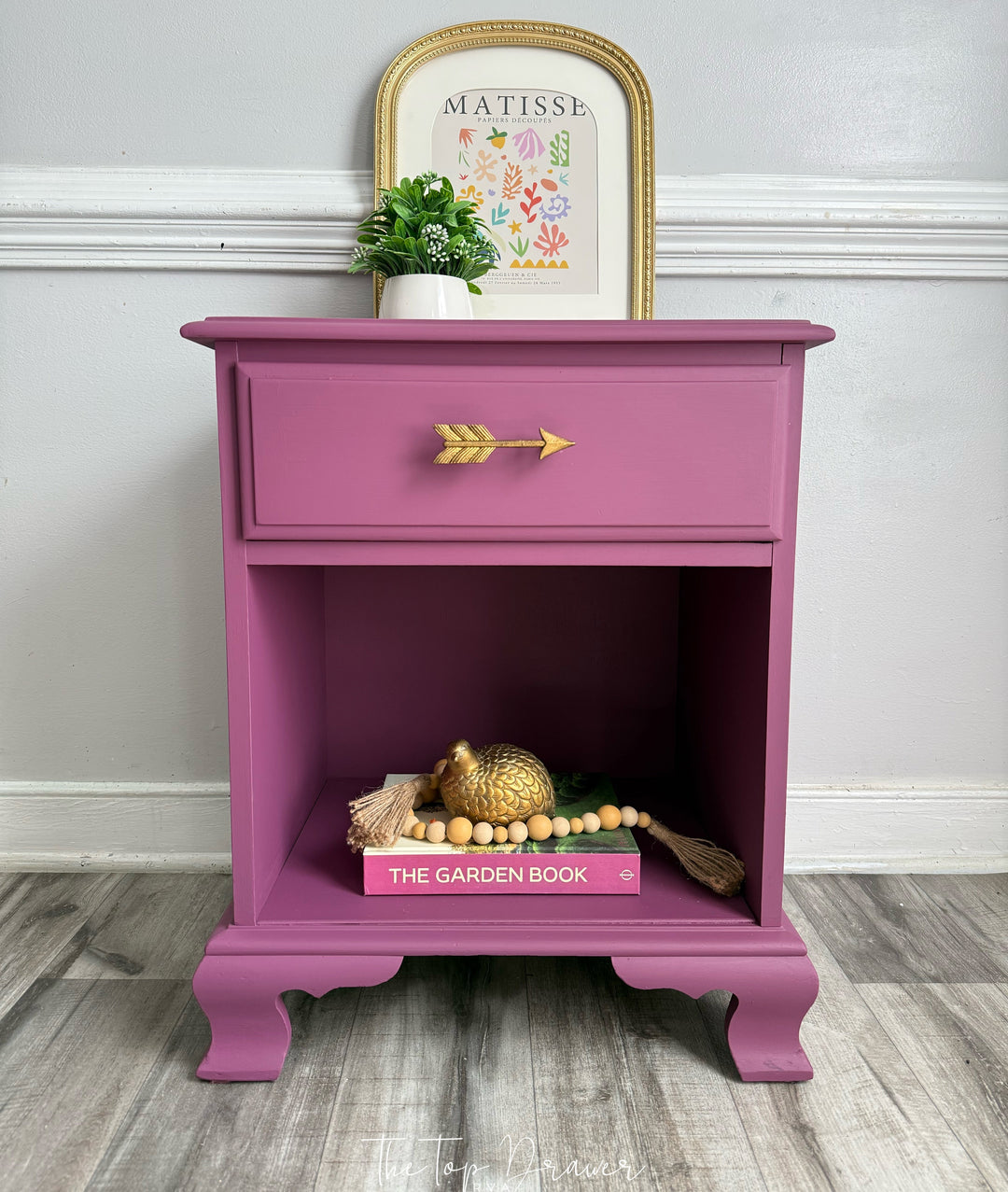 A magenta-painted nightstand with a single drawer and open shelf, styled with a small potted plant, a framed print, and decorative items on a gray wood floor against a white wall.