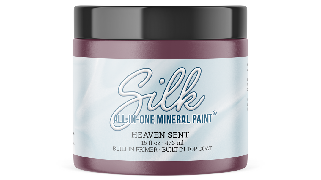A jar of Silk All-In-One Mineral Paint in the scent Heaven Sent, featuring a deep plum purple color visible through the clear jar, a black lid, and a white label with blue text.