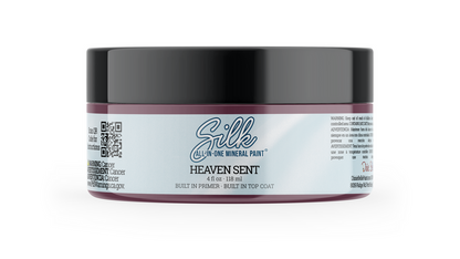 A jar of Silk All-In-One Mineral Paint in the shade Heaven Sent, featuring a black lid and a white label with purple accents, set against a plain white background.