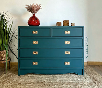A teal blue wooden chest of drawers with brass handles, styled in a room setting with a red vase on top, a potted plant on the left, and decorative blocks on the right. A vertical text overlay reads 'ATELIER 13LX'.