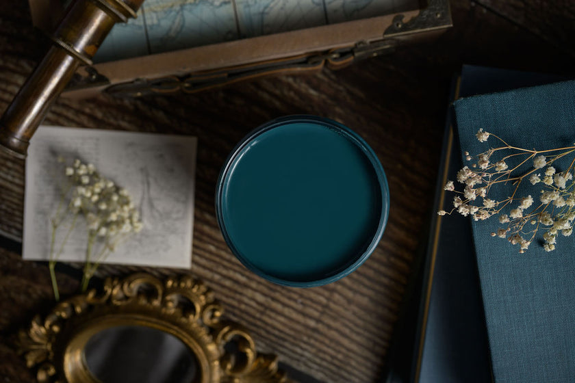 Teal heirloom paint container on a wooden surface with dried flowers and an ornate mirror.
