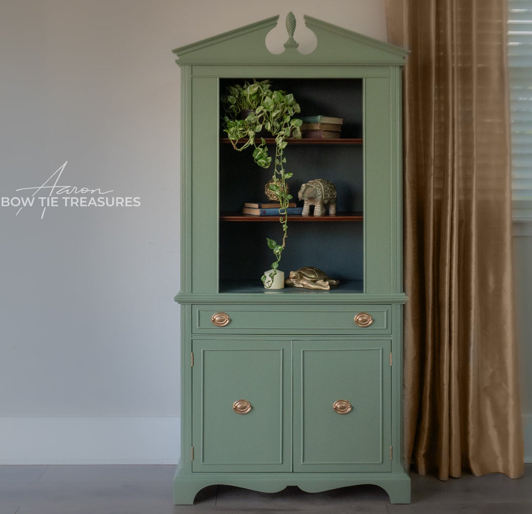 A tall, mint green wooden hutch cabinet with a glass-front upper section and two lower drawers, styled with books, a plant, and decorative objects. The cabinet features brass handles and is set against a neutral wall with a brown curtain on the right.