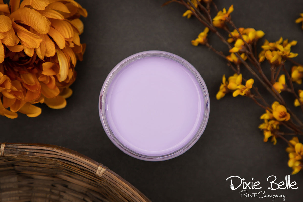dixie Belle Paint color Luxy Lavender container on a dark surface with yellow flowers and a wicker basket.