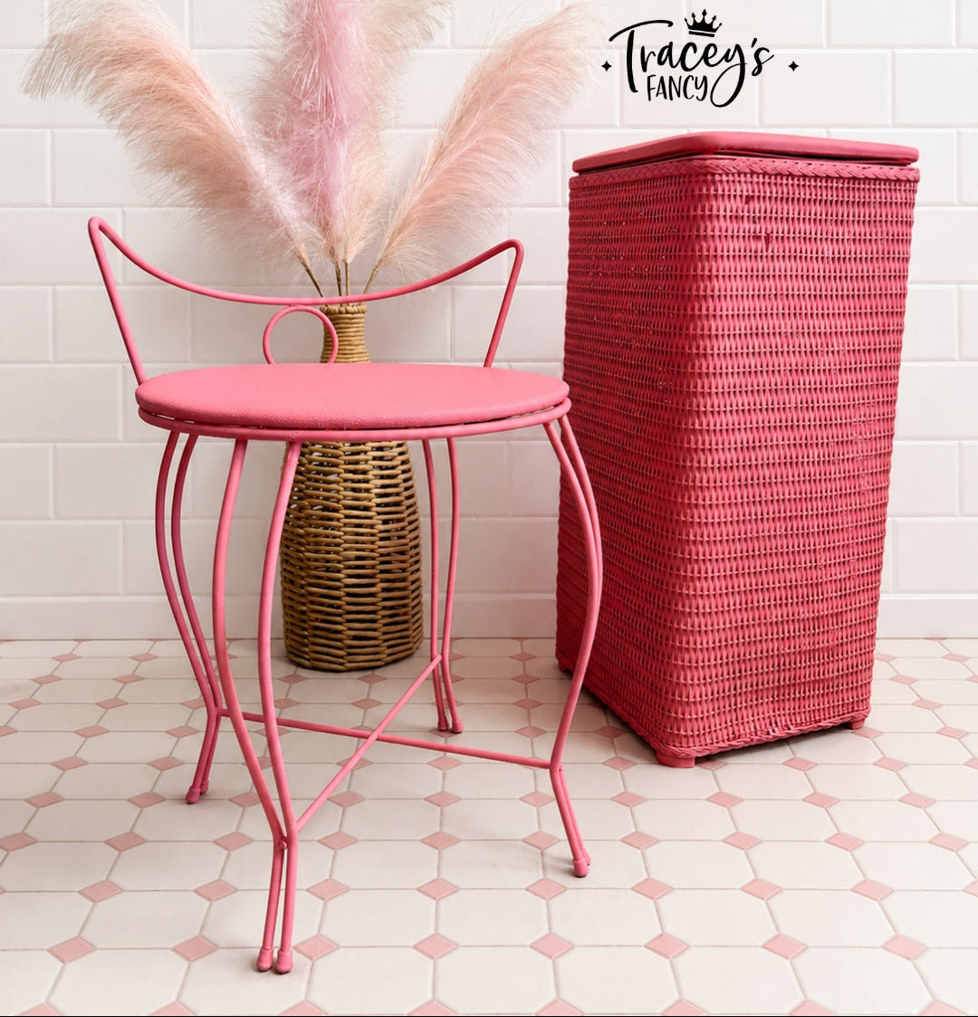 A pink metal side stool with a round cushioned seat and curved backrest, shown beside a tall woven red storage bin. A wicker vase with pink pampas grass sits behind the stool against a white tiled wall, and the brand name 'Tracey's Fancy' appears in the top right.