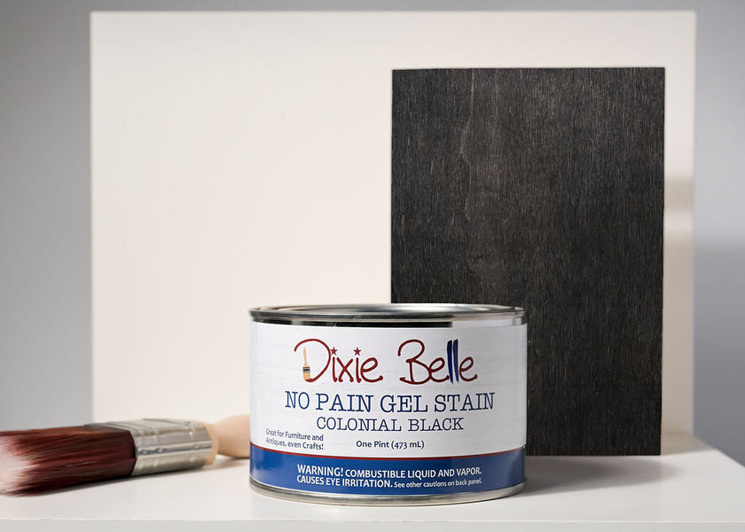 A can of Dixie Belle No Pain Gel Stain in Colonial Black, shown in front of a black rectangular sample board on a white surface, with a paintbrush resting to the left.