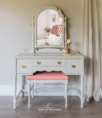 A white painted wooden vanity table with an arched mirror, three drawers with gold-tone handles, and a matching bench with a red-and-white gingham cushion, styled in a bedroom setting.