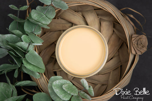 Dixie Belle Paint color Rebel Yellow Jar in a woven basket with greenery on a dark background, featuring the Dixie Belle Paint Company logo.
