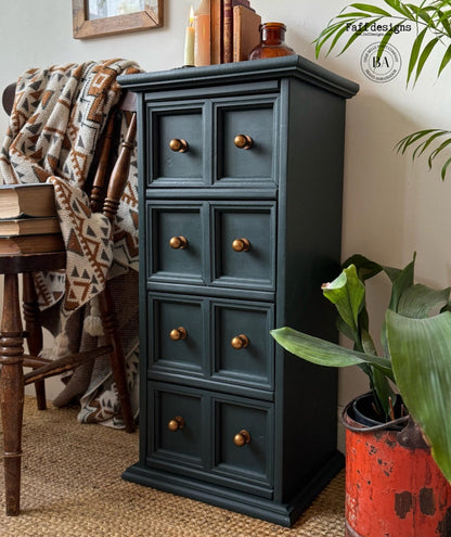 Dark green cabinet with gold knobs in a home setting with a plant and books.