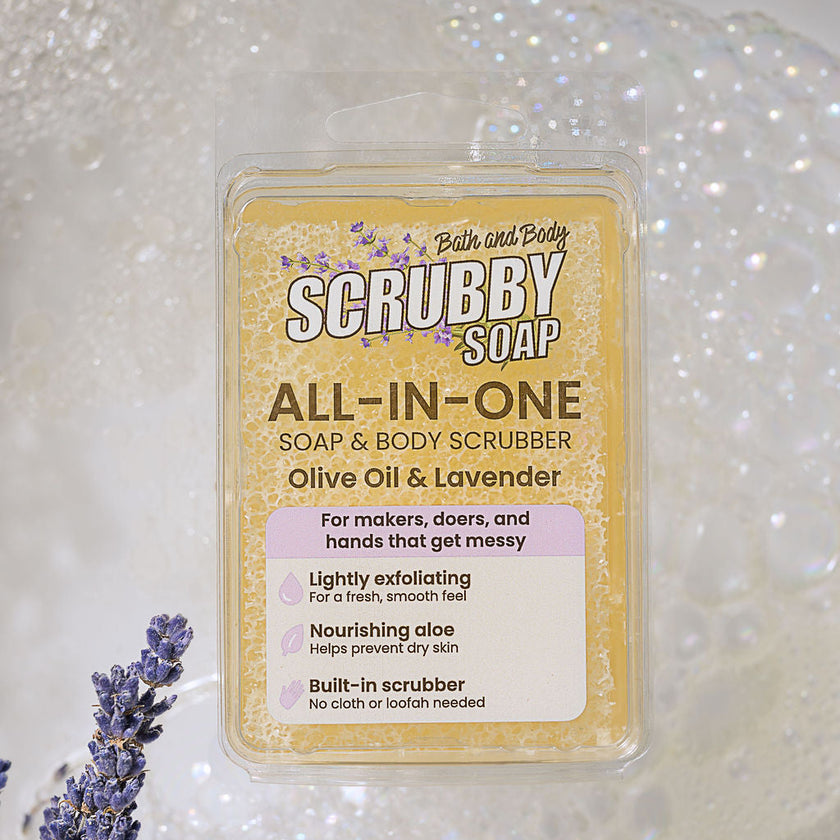 A clear plastic tub of Bath and Body Scrubby Soap, All-In-One, Olive Oil & Lavender, shown against a sparkling white background with decorative lavender sprigs.