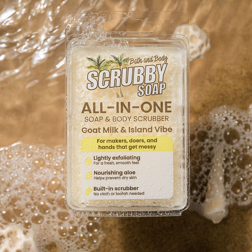 A clear plastic tub of Bath and Body Scrubby Soap, 'Goat Milk & Island Vibe', surrounded by soapy foam on a warm, sandy brown background.