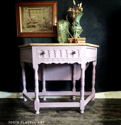 A vintage-style wooden writing desk painted in a dusty lavender-pink finish, featuring a light wood top, a single drawer with decorative knobs, and turned legs. It is styled with a green ceramic vase and books on top, set against a dark background.