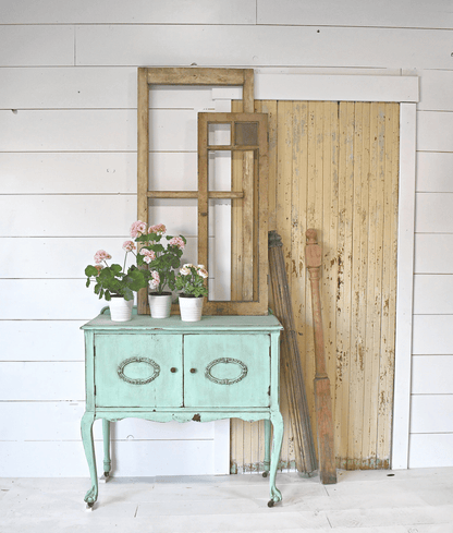 Vintage-style green cabinet with floral arrangements, wooden mirror, and planks against a white wall.