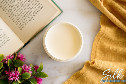 An open jar of creamy off-white Sunkissed Paint on a light marble surface, styled with an open book, pink flowers, a yellow cloth, and a 'Silk All-In-One Mineral Skin' logo in the corner.
