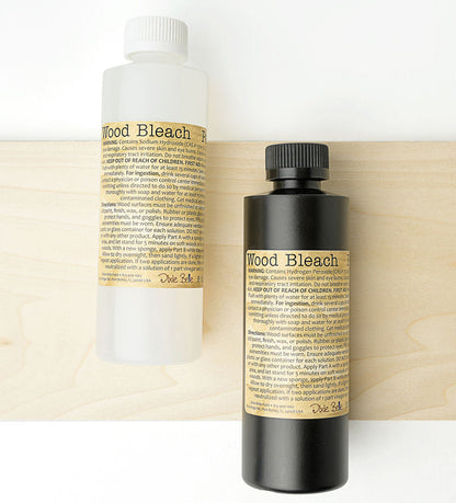 Two bottles of Dixie Belle Wood Bleach, one in a translucent white bottle and one in a black bottle, both with tan labels, set against a light wood panel background.