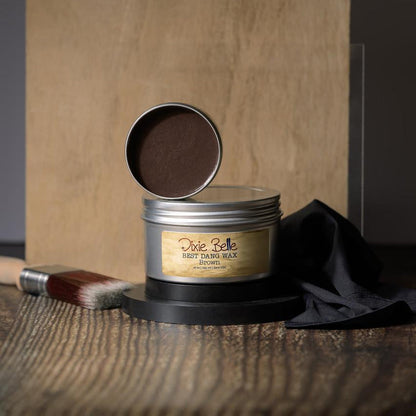 A tin of Dixie Belle Wax in Brown, shown open with the lid resting on top, displayed on a wooden surface with a black cloth and a paintbrush nearby.