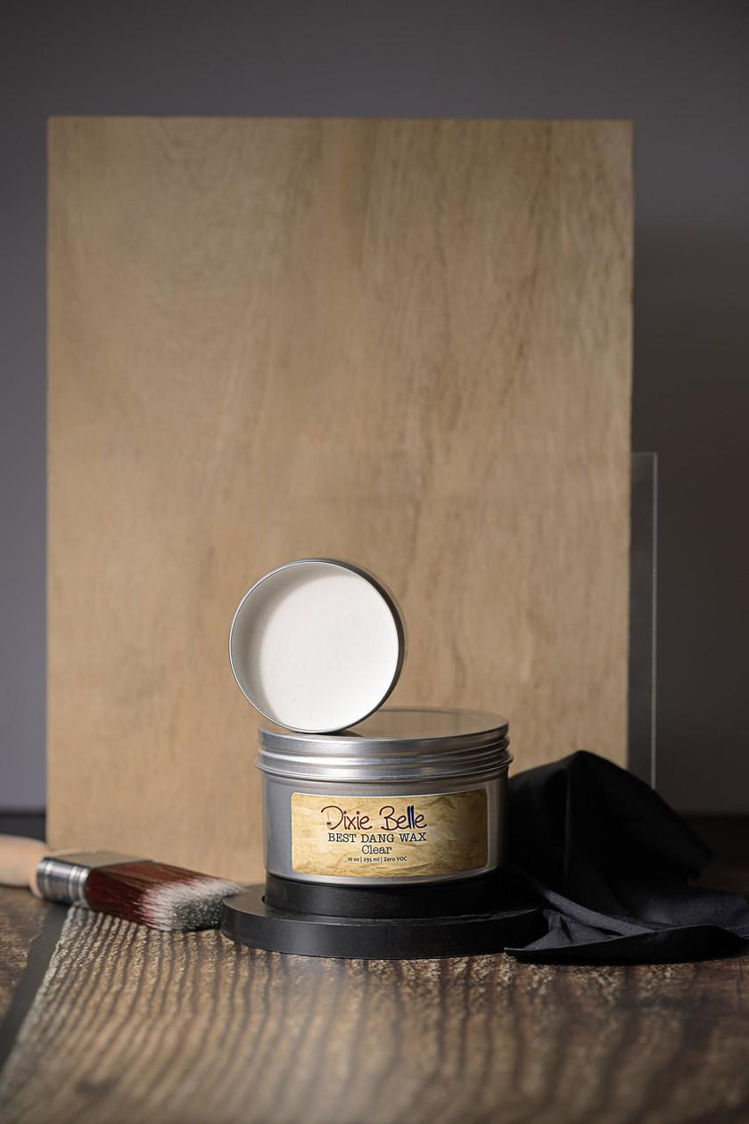 Small tin with a lid on a wooden surface next to a brush and black cloth, with a brown wood plank in the background.