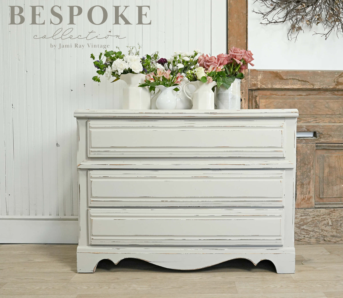 Vintage-style chest of drawers with floral arrangements on top, against a wooden wall.