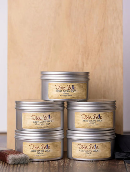 Stack of Dixie Belle wax tins on a wooden surface with a brush in the foreground.