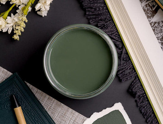 A round clear glass jar filled with muted sage green cream or balm, photographed from above on a dark surface with decorative props including white flowers, a notebook, and an open book.