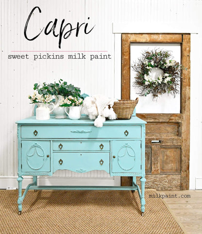 A turquoise painted wooden console table styled with white flowers, a white plush toy, and a wicker basket, set against a white paneled wall with a rustic wooden door and wreath. Text on the image reads 'Capri sweet pickins milk paint'.