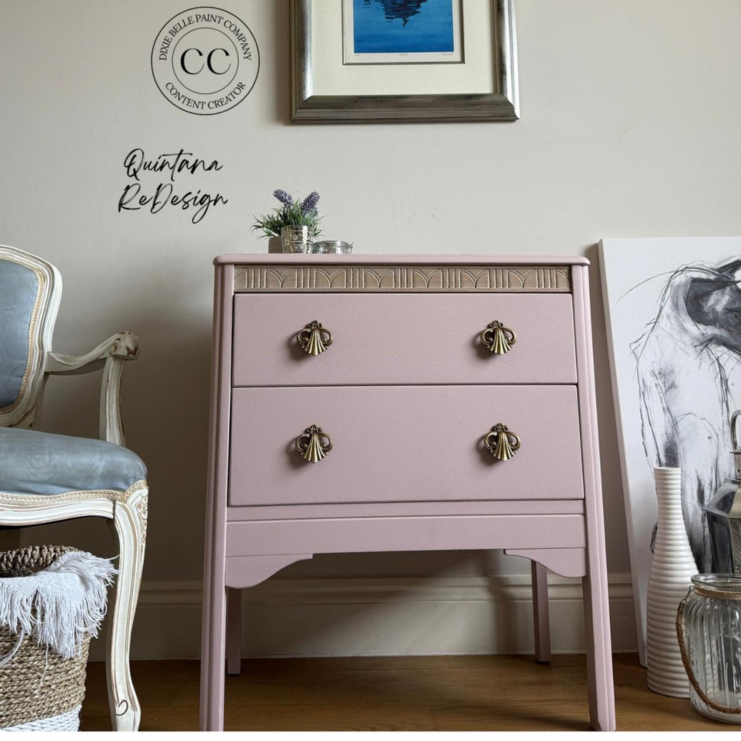 A dusty pink two-drawer nightstand with decorative brass handles and a carved gold-toned trim, styled in a cozy room setting next to a blue upholstered chair.