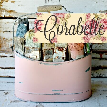 A distressed pink metal paint bucket filled with paintbrushes, featuring a floral banner that reads 'Corabelle' across the front, set against a rustic white wooden plank background.