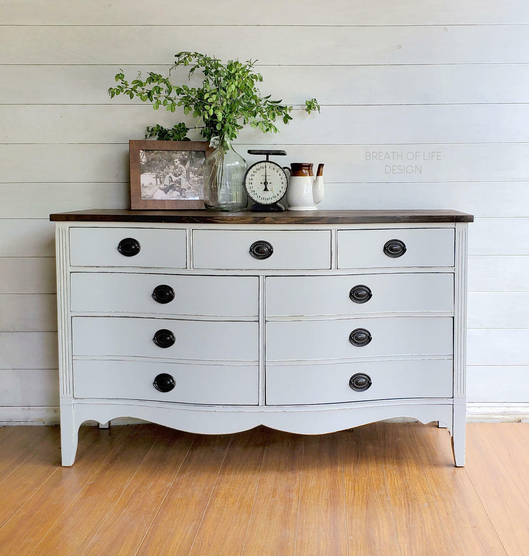 A white painted wooden dresser with a dark brown top, featuring multiple drawers with black knobs, styled with home decor items including a vase of greenery, a clock, and small ceramic containers against a white shiplap wall and wood floor.