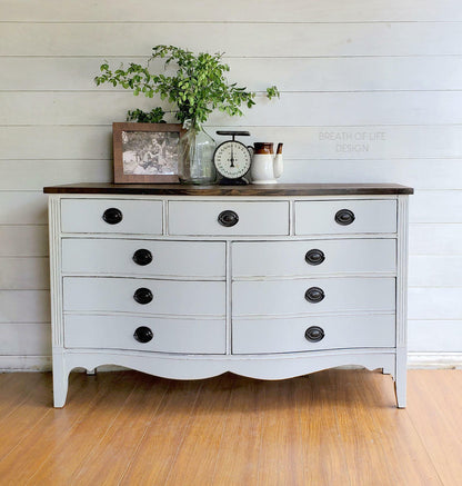A white painted wooden dresser with a dark brown top, featuring multiple drawers with black knobs, styled with home decor items including a vase of greenery, a clock, and small ceramic containers against a white shiplap wall and wood floor.